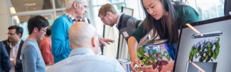 A female student holds a plant that has been used in her research project, and is describing it to an attentive man who is seated.